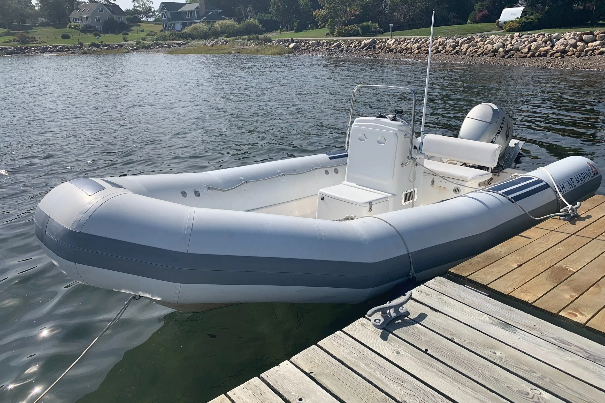 A white and gray inflatable boat docked at a wooden pier on a calm body of water with houses and greenery in the background.