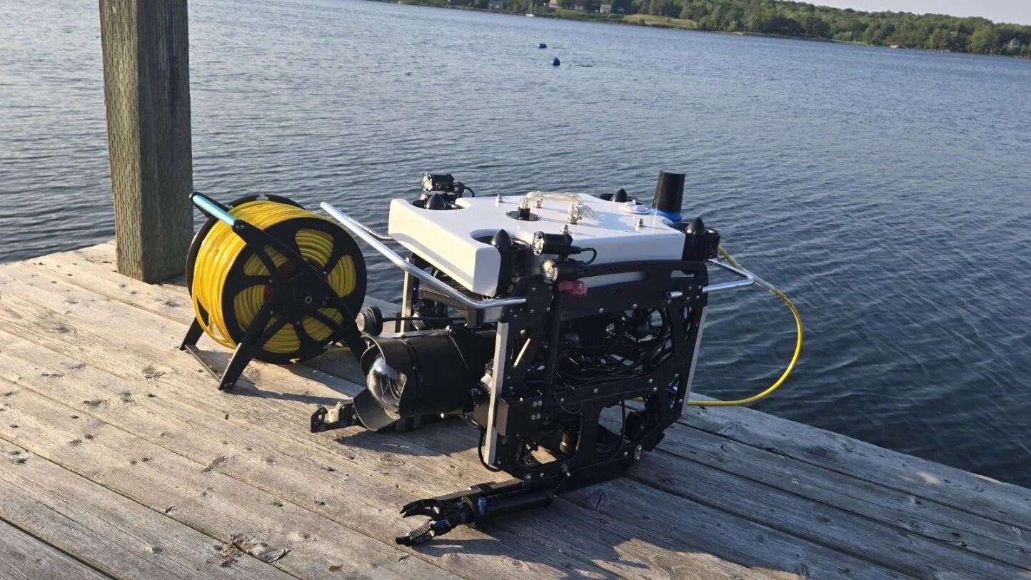 Remotely operated underwater drone with a yellow cable reel on a wooden dock by a lake, with water and trees in the background.