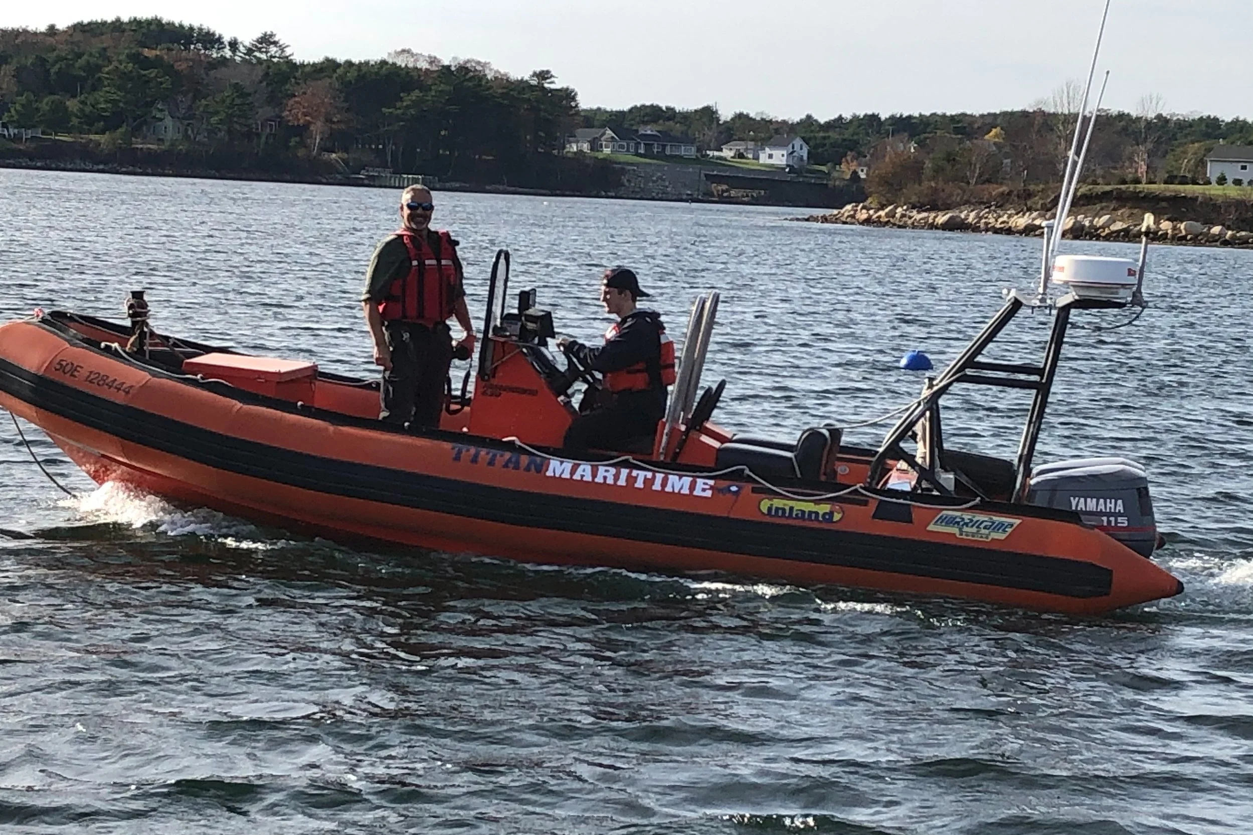 Two people on an orange Titan maritime rescue boat on a body of water, with houses and trees in the background.