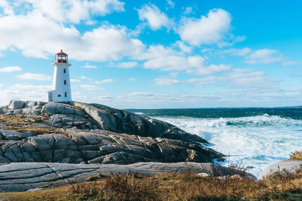 A lighthouse on rocky coast near the ocean, with waves crashing and a partly cloudy blue sky.