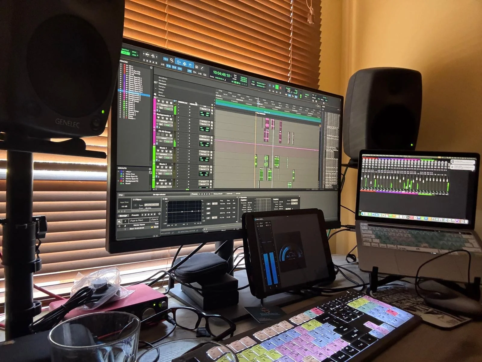 A music production workspace with multiple screens showing audio editing and mixing software, studio monitors, a keyboard with colorful keys, and other electronic equipment.