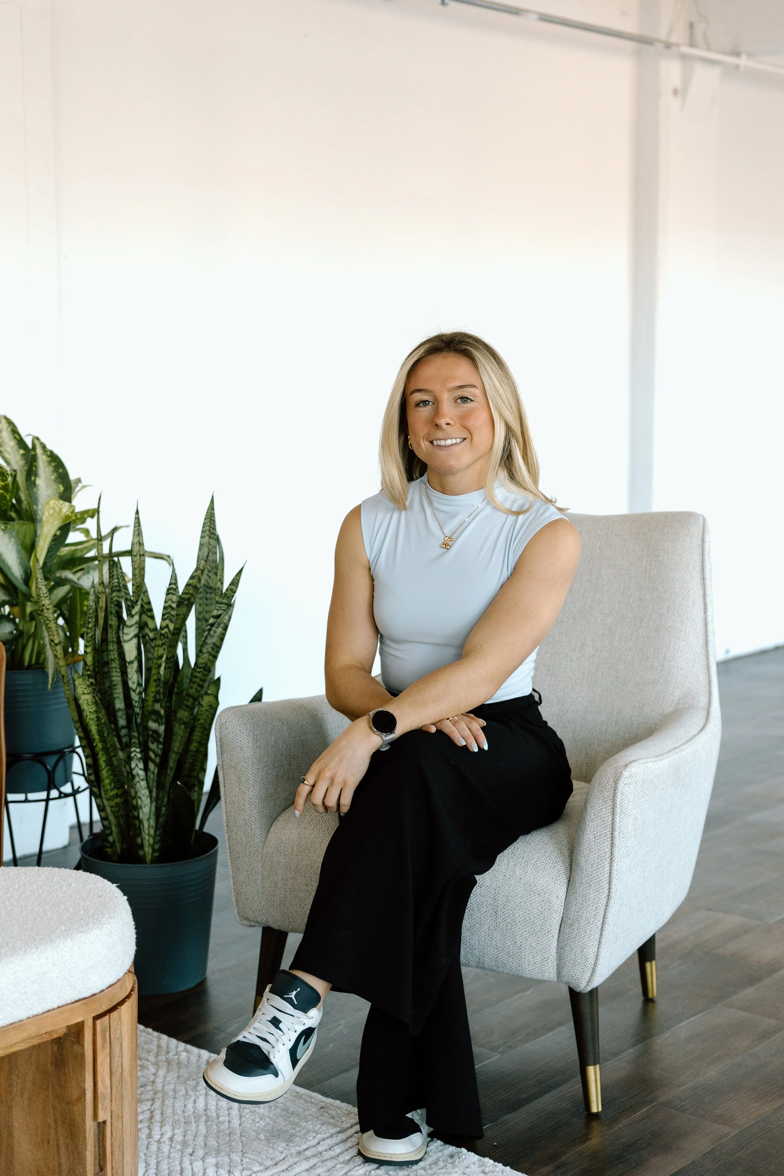 A young woman in a sleeveless white top and black pants sitting on a beige armchair with plants nearby in a well-lit room.
