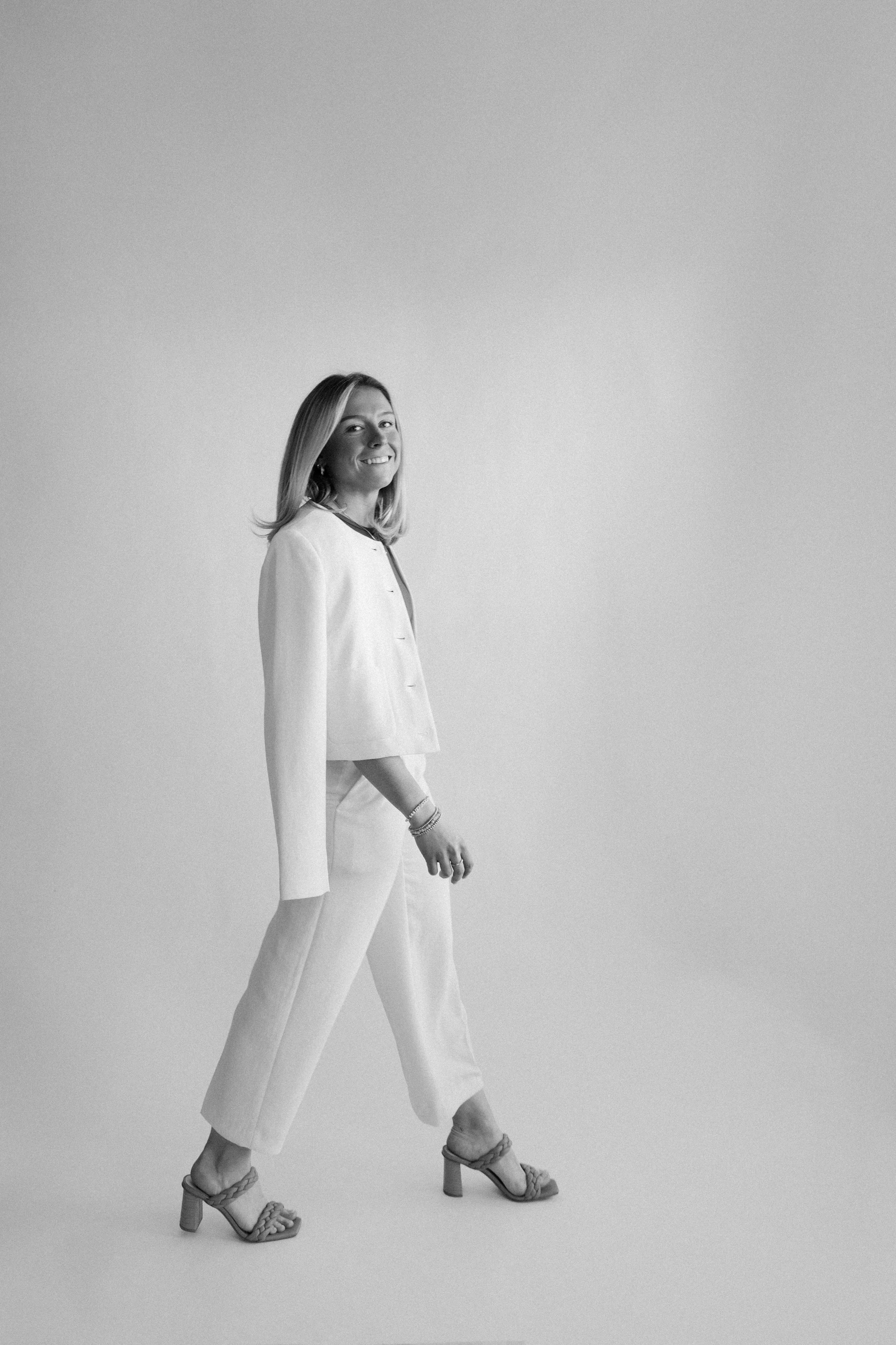 Black and white photo of a woman in a pantsuit and high heels standing sideways, smiling and looking at the camera against a plain background.