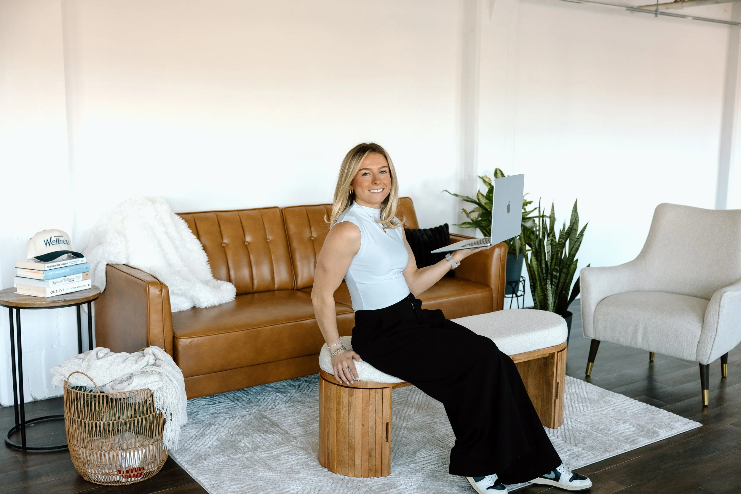 A smiling woman sitting on a small wooden stool, holding a laptop, in a modern living room with a tan leather sofa, white armchair, green plants, and a side table with books.