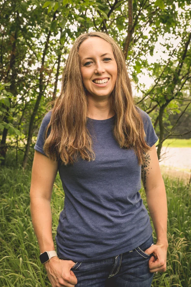 A woman with long curly hair smiling outdoors in a green, wooded area, wearing a blue t-shirt and jeans.