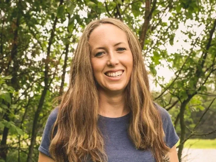 A woman with long, wavy brown hair smiling outdoors among green trees.