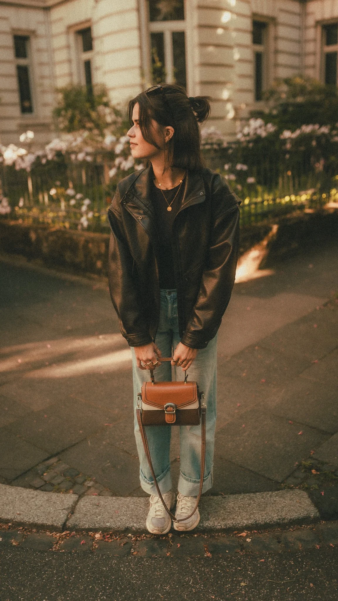 Eine junge Frau mit dunklen Haaren, die eine Lederjacke und Jeans trägt, steht nachts auf der Straße vor einem Haus mit Blumen im Hintergrund. Sie hält eine braune Handtasche.