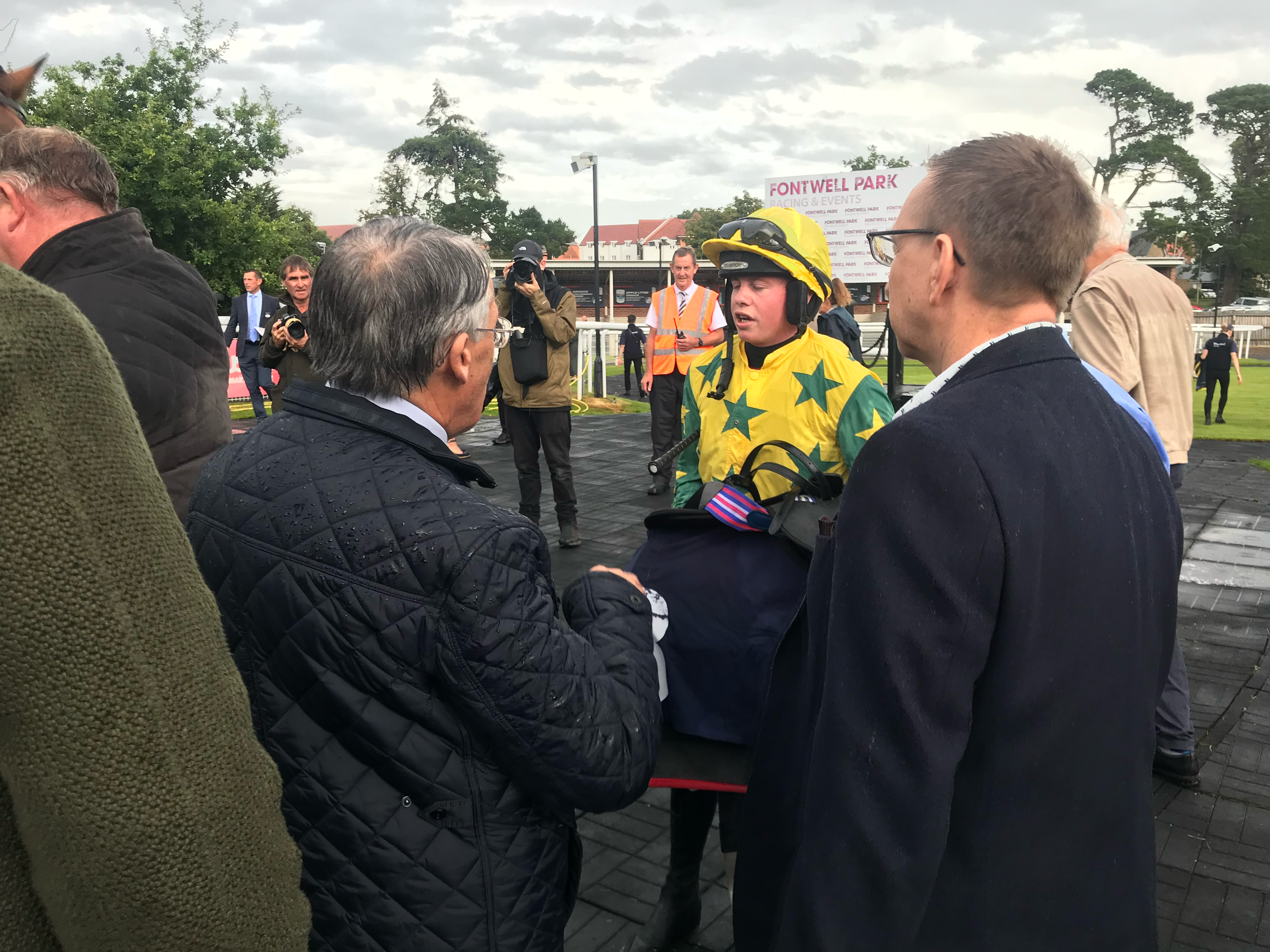 Group of people talking outdoors near a fence, with one person wearing a yellow jacket with green stars, and some individuals holding cameras.