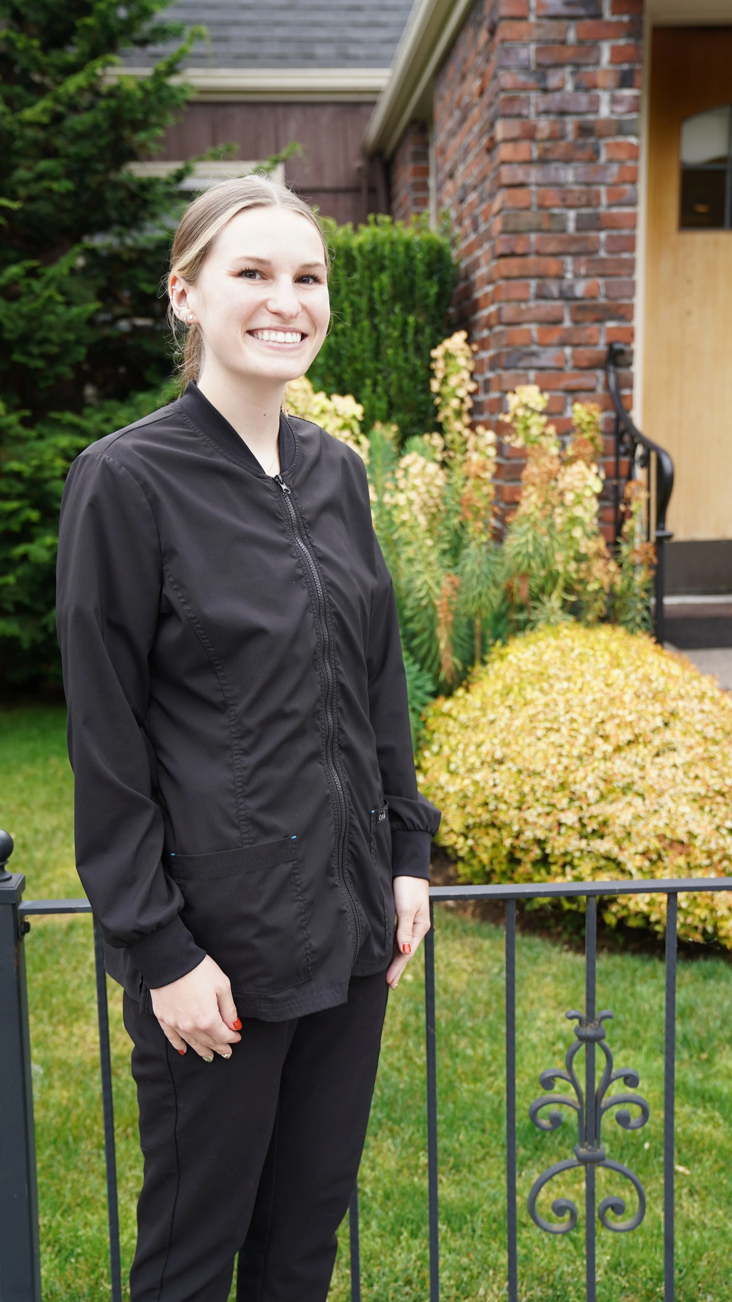 A young woman with blonde hair smiling and wearing a black jacket standing outdoors in front of a garden with green bushes and yellow plants, near a brick house with stairs.