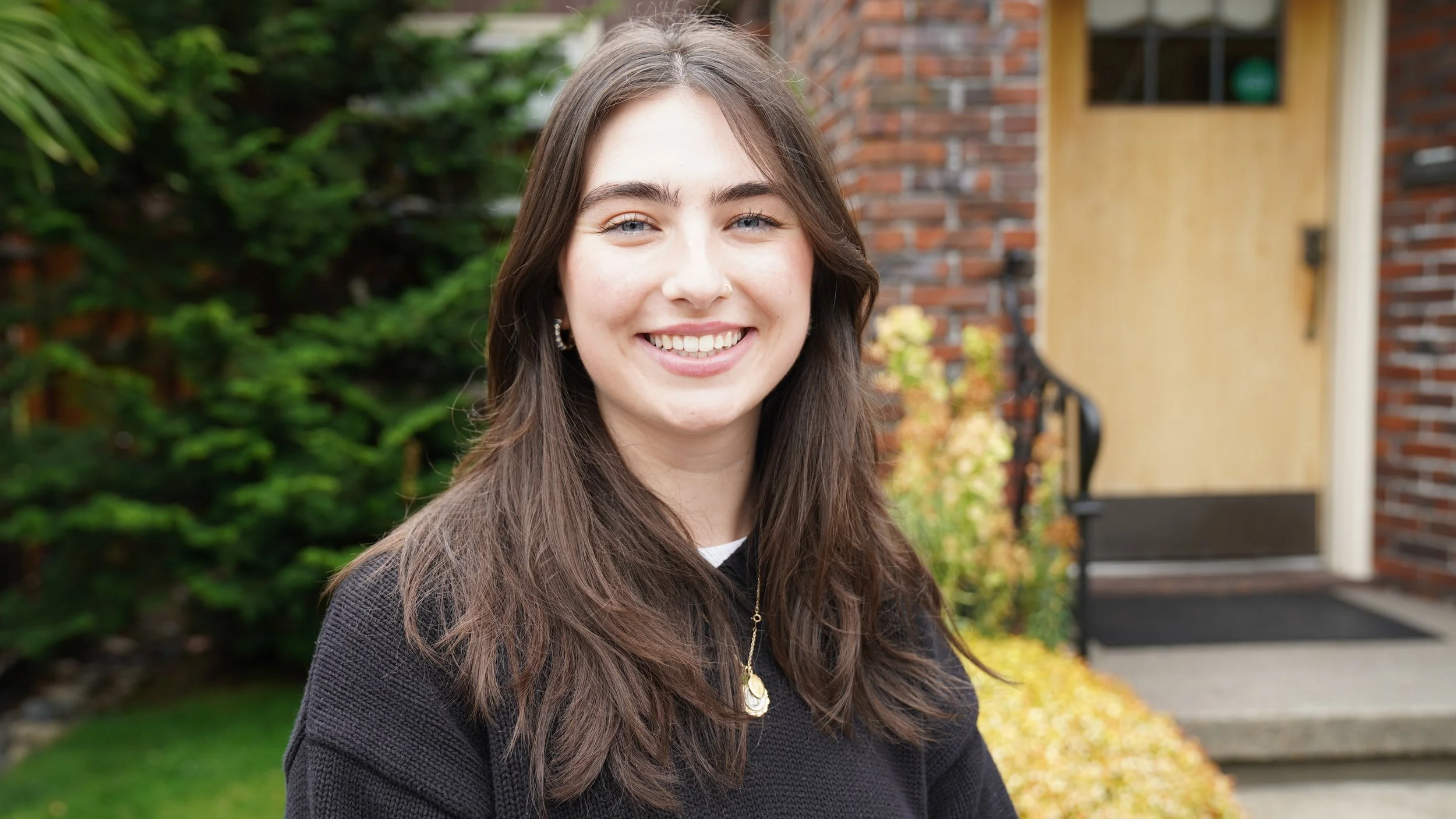 Smiling young woman with long brown hair and blue eyes standing outside in front of a brick house with an open yellow door, green bushes, and autumn leaves on the ground.