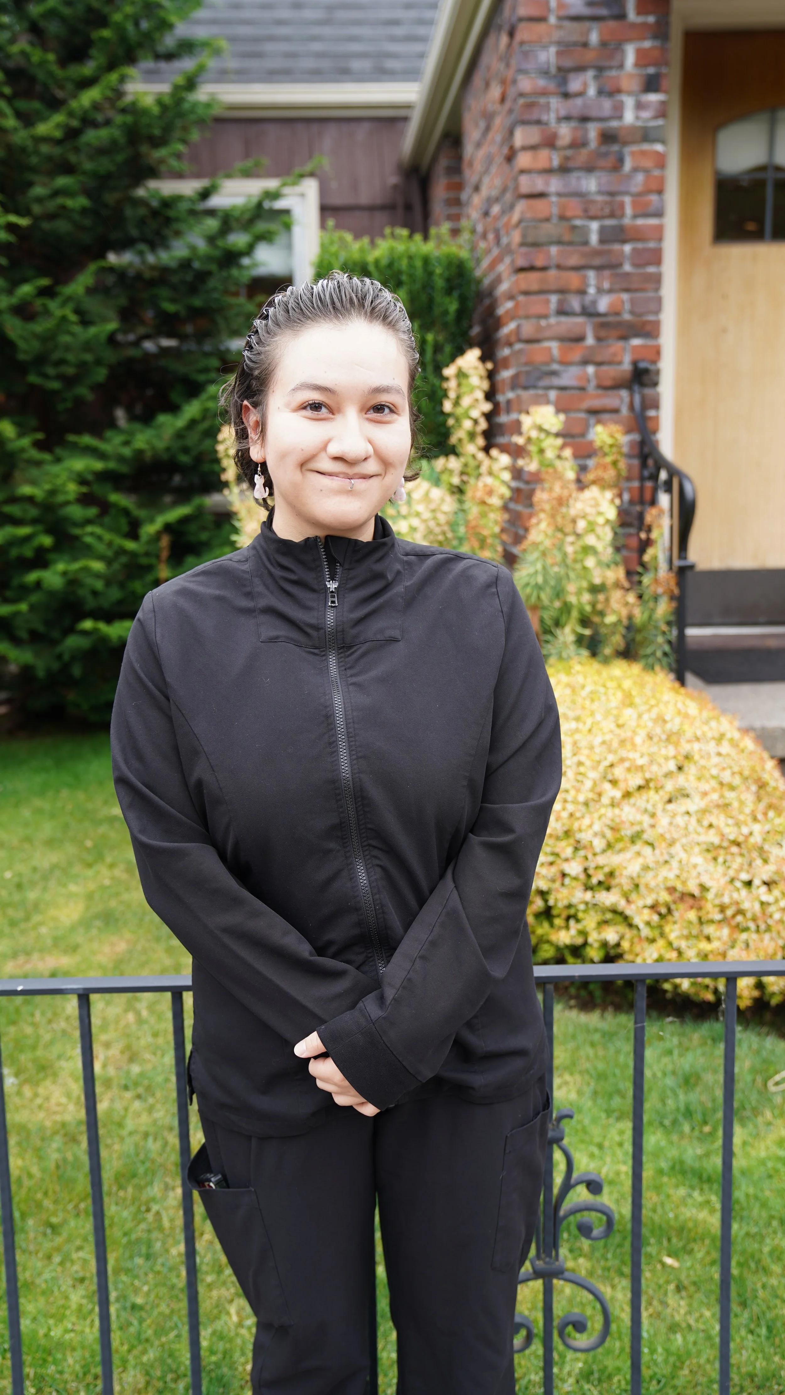 A woman with short dark hair and earrings standing outdoors, wearing a black zip-up jacket and black pants, in front of a garden with greenery and a brick house.