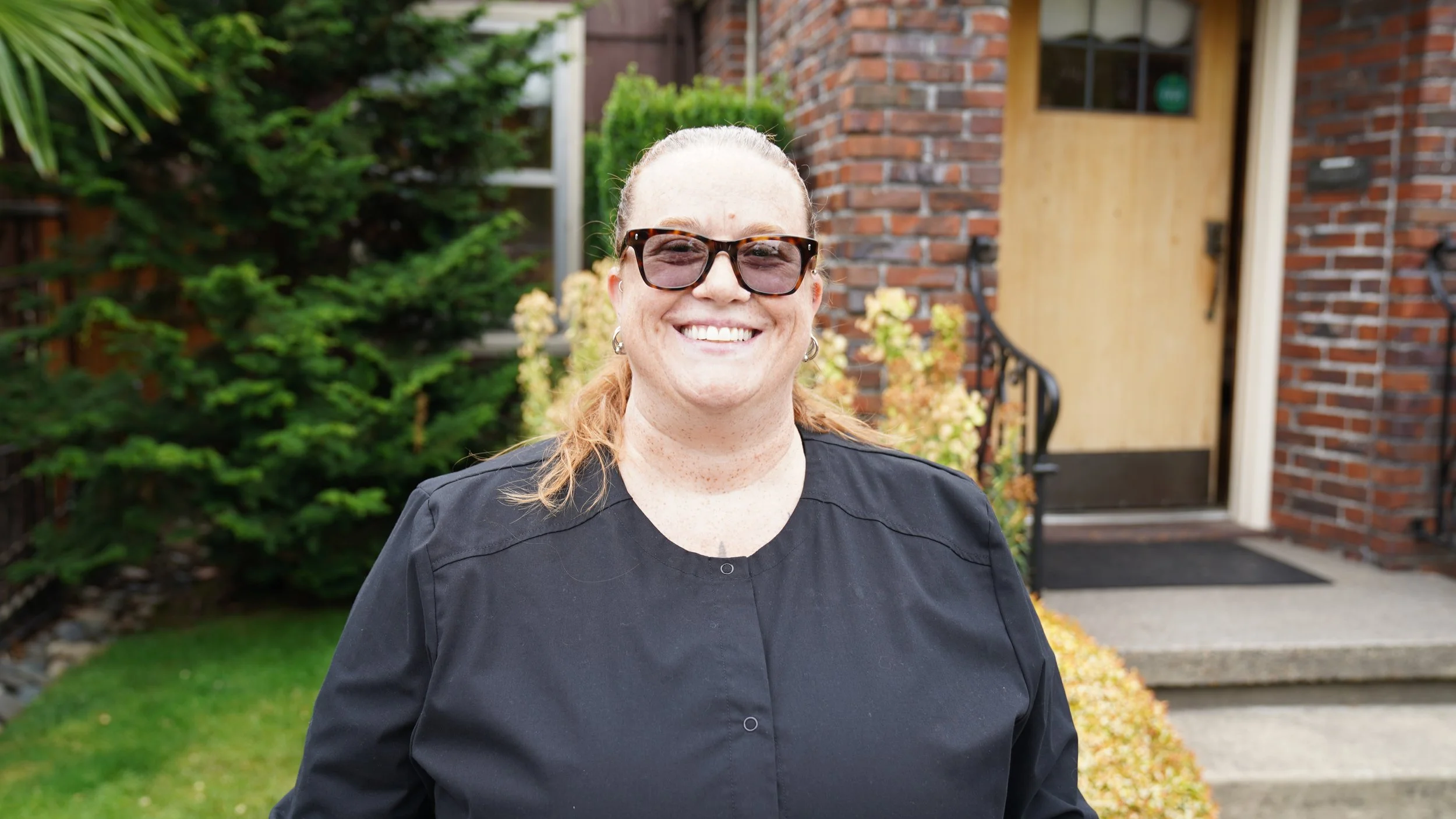 A woman wearing sunglasses and a black top smiling outdoors in front of a brick house with a wooden door and garden plants.