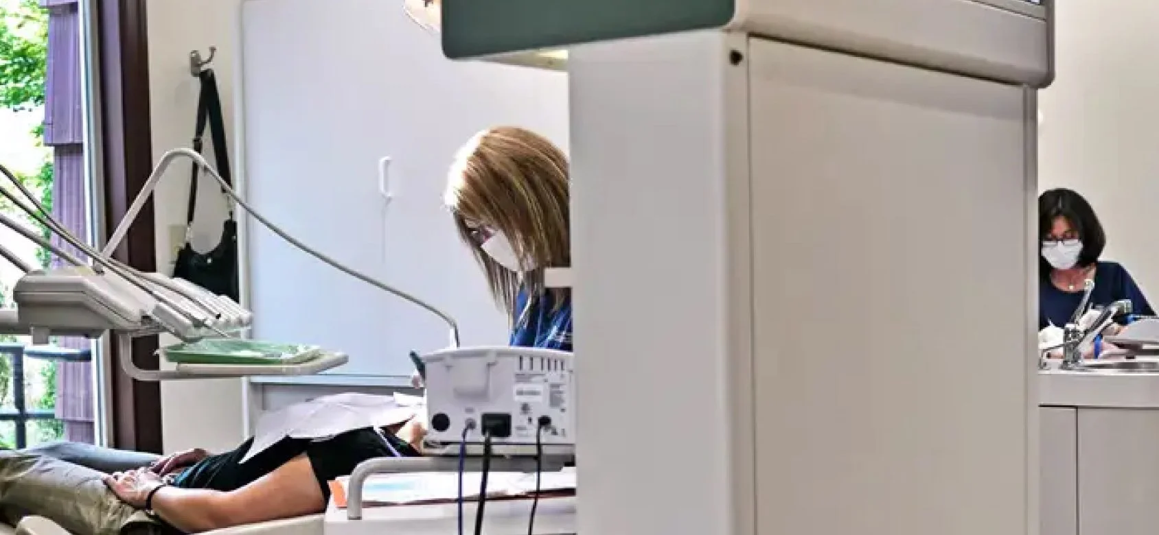 A woman lying on a hospital bed talking to a healthcare worker sitting at a desk, both wearing face masks, in a medical clinic.