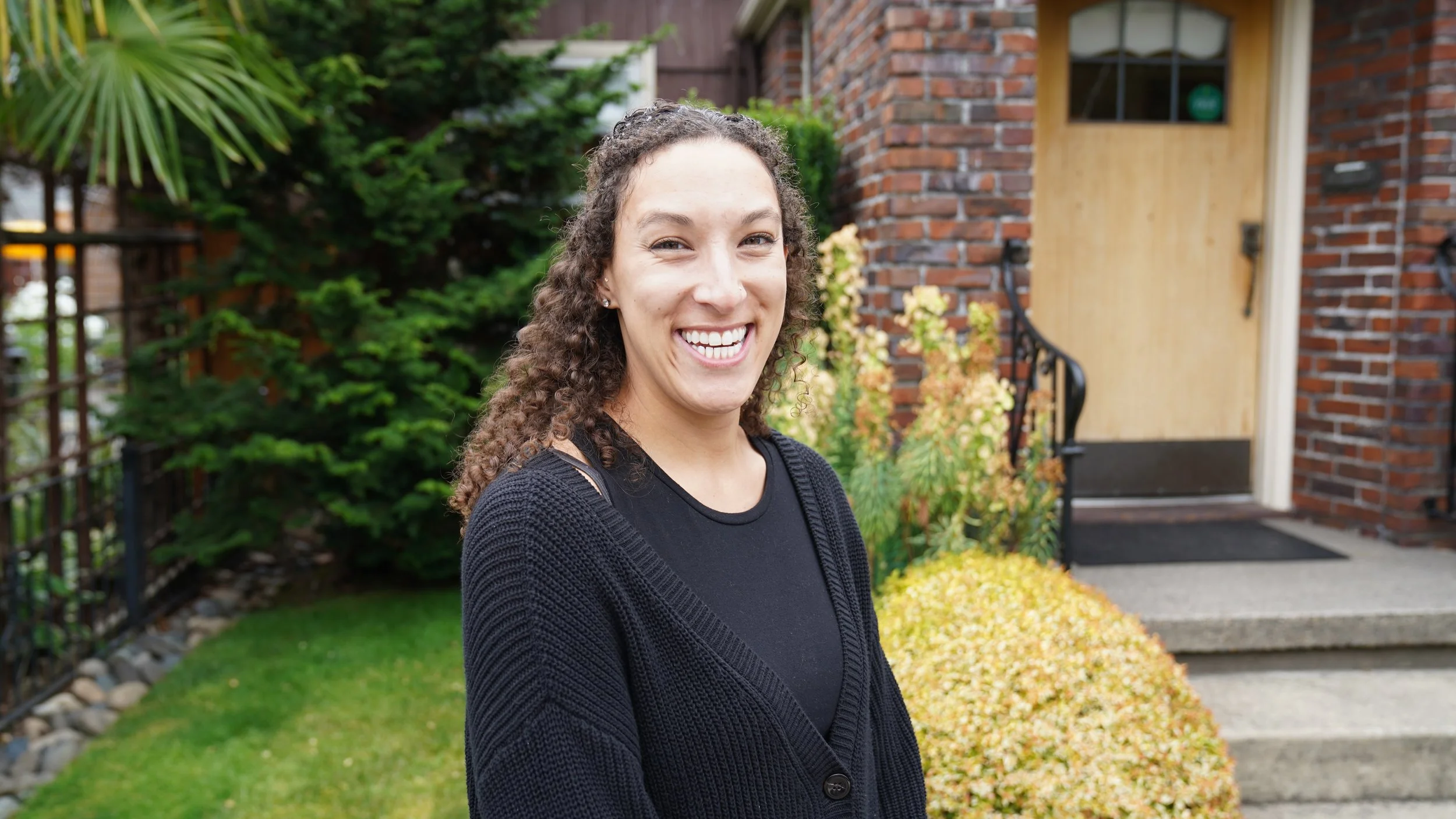 A woman with curly hair and a black sweater smiling outside in front of a brick house with a wooden door, greenery, and yellow shrubs.