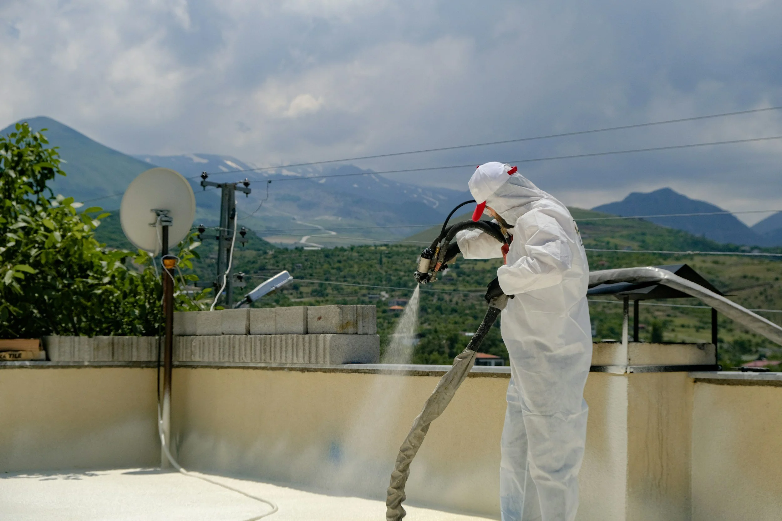 Person in full protective suit with hood and mask using a spray hose outdoors on a rooftop, with trees, mountains, and a cloudy sky in the background.