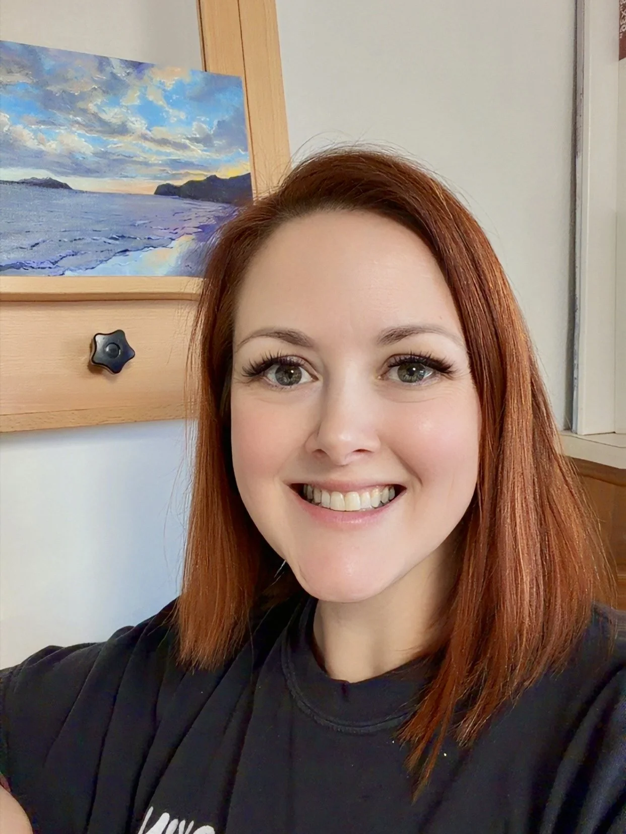 A woman with shoulder-length red hair smiling in front of a painting of a seascape with rocks and clouds.