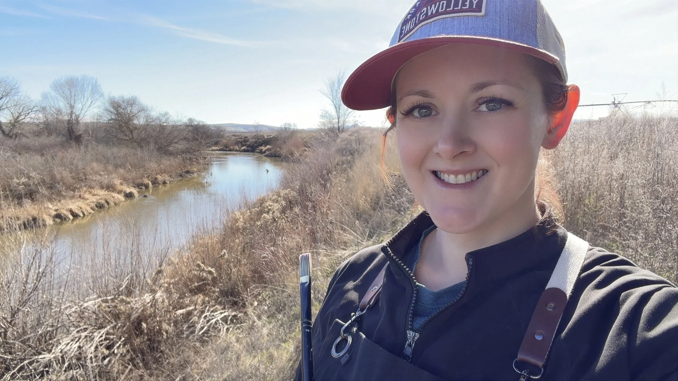 An artist smiling outdoors near a small river or creek with dry bushes and trees in the background, plein air painting.