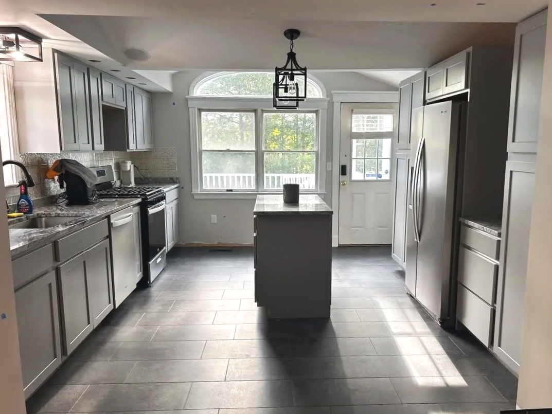 Modern kitchen with gray cabinets, black countertop, stainless steel appliances, a central island, large window, and ceramic tile flooring.