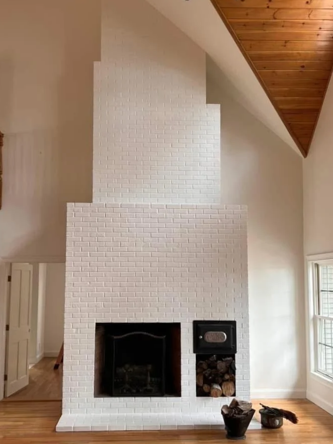 White brick fireplace in a living room with a wooden ceiling and hardwood floor, featuring a wood storage area, a black cast iron firebox, and a window to the right.