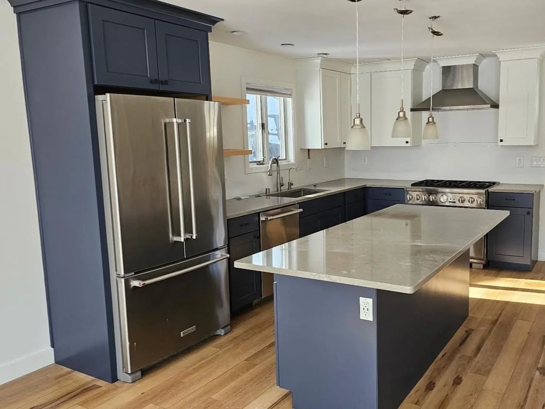 Modern kitchen with blue cabinets, white upper cabinets, stainless steel refrigerator, countertop island, stainless steel oven range, and hanging pendant lights.