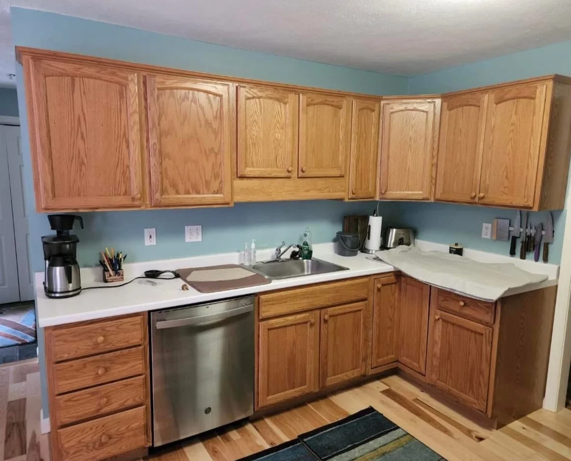 Kitchen with wooden cabinets, stainless steel dishwasher, coffee maker, cutting board, and various kitchen utensils.