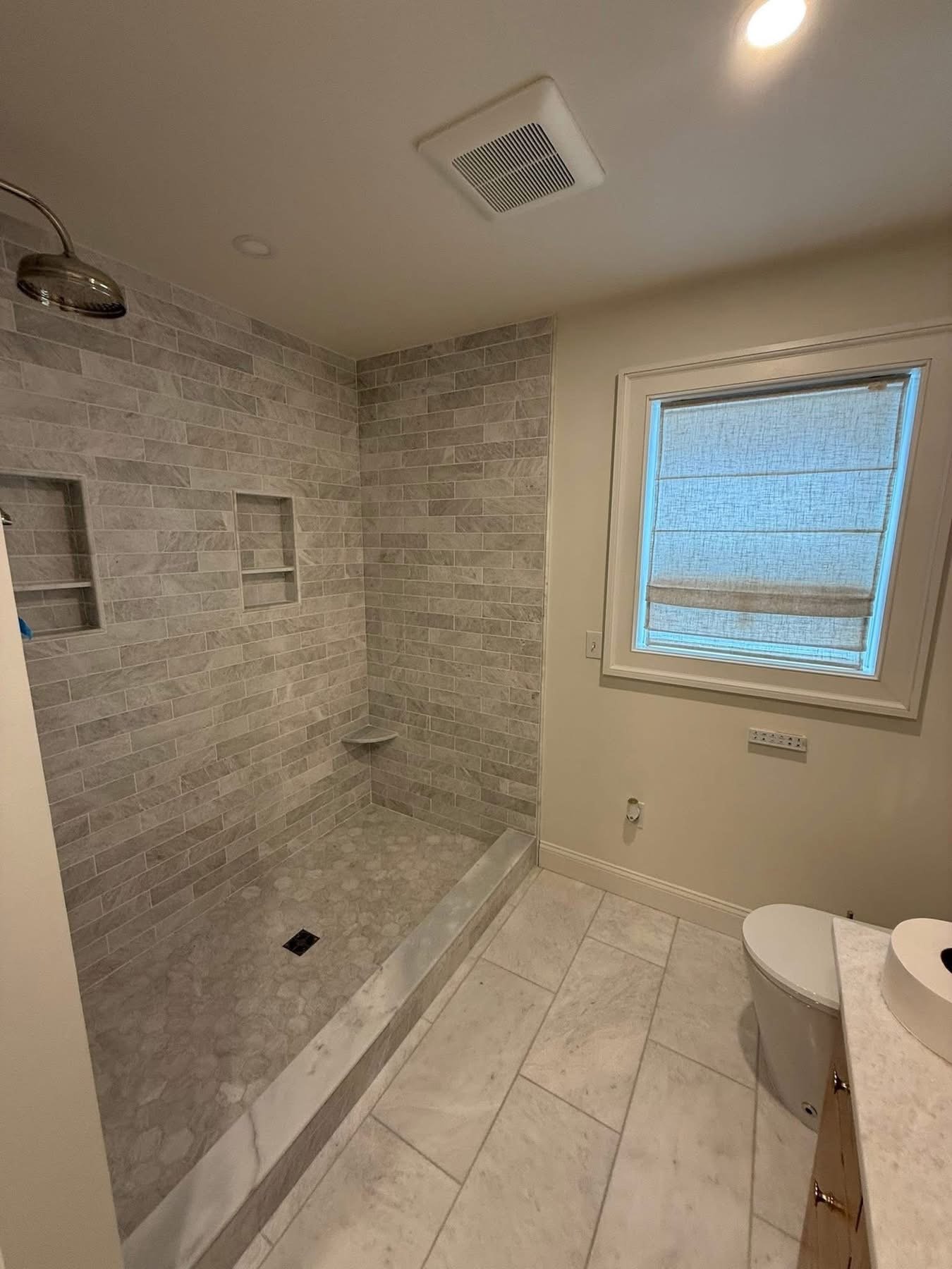 A bathroom with a tiled walk-in shower, a window with a roller shade, a toilet, and a vanity with a vessel sink.