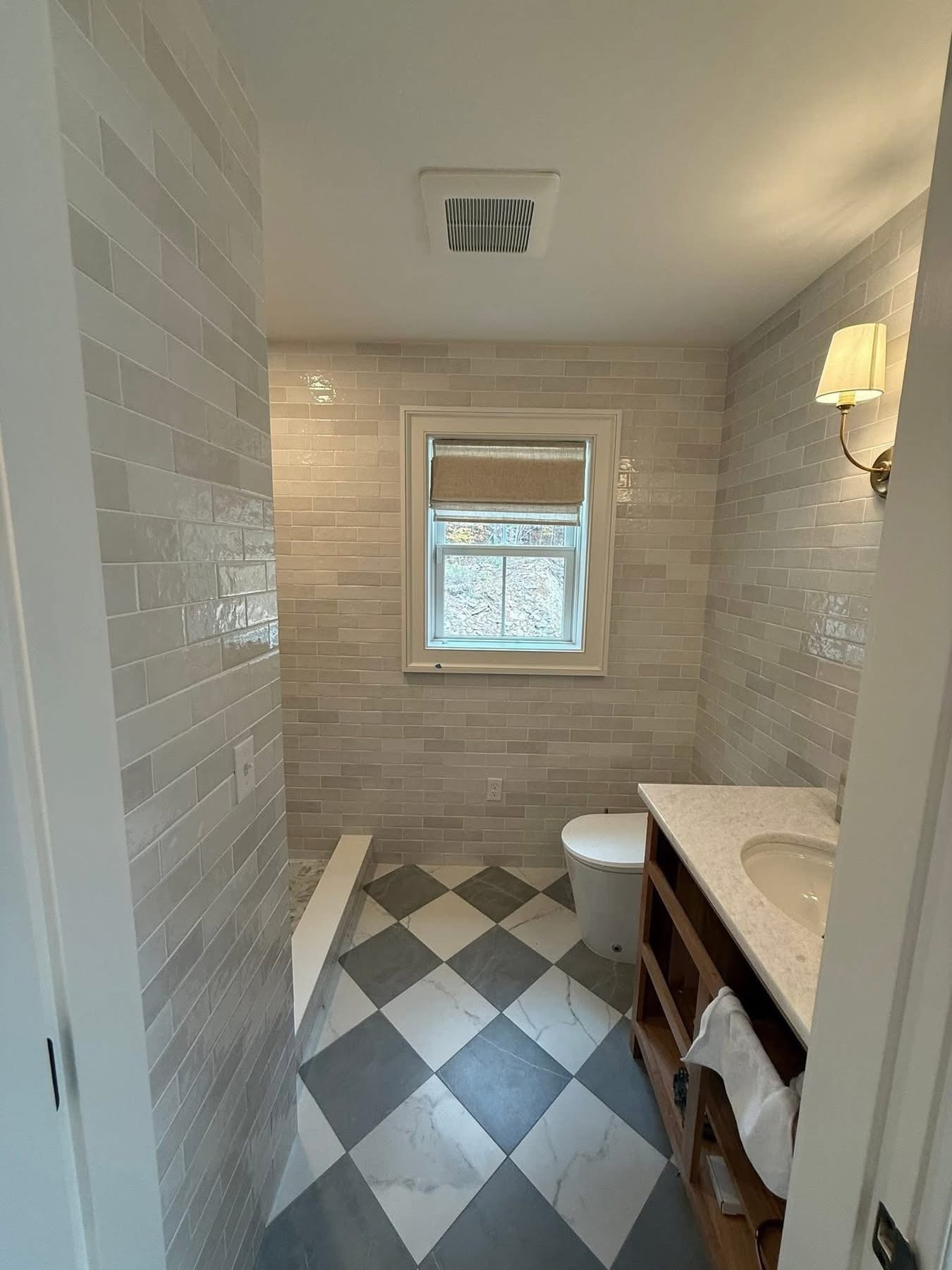 Bathroom with checkered black and white floor tiles, beige brick walls, a window with a beige shade, a wall-mounted lamp, a toilet, and a wooden vanity with a countertop.