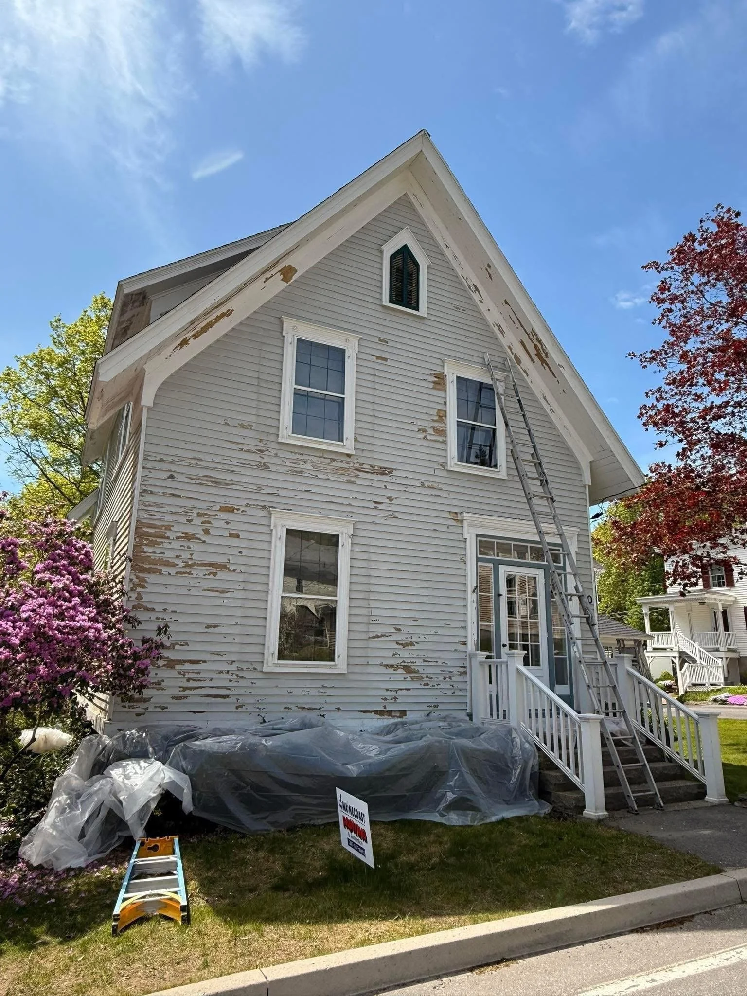 A two-story house with peeling white paint on the exterior, a small front porch with stairs, a tall ladder leaning against the house, and a construction sign on the lawn indicating roof work.