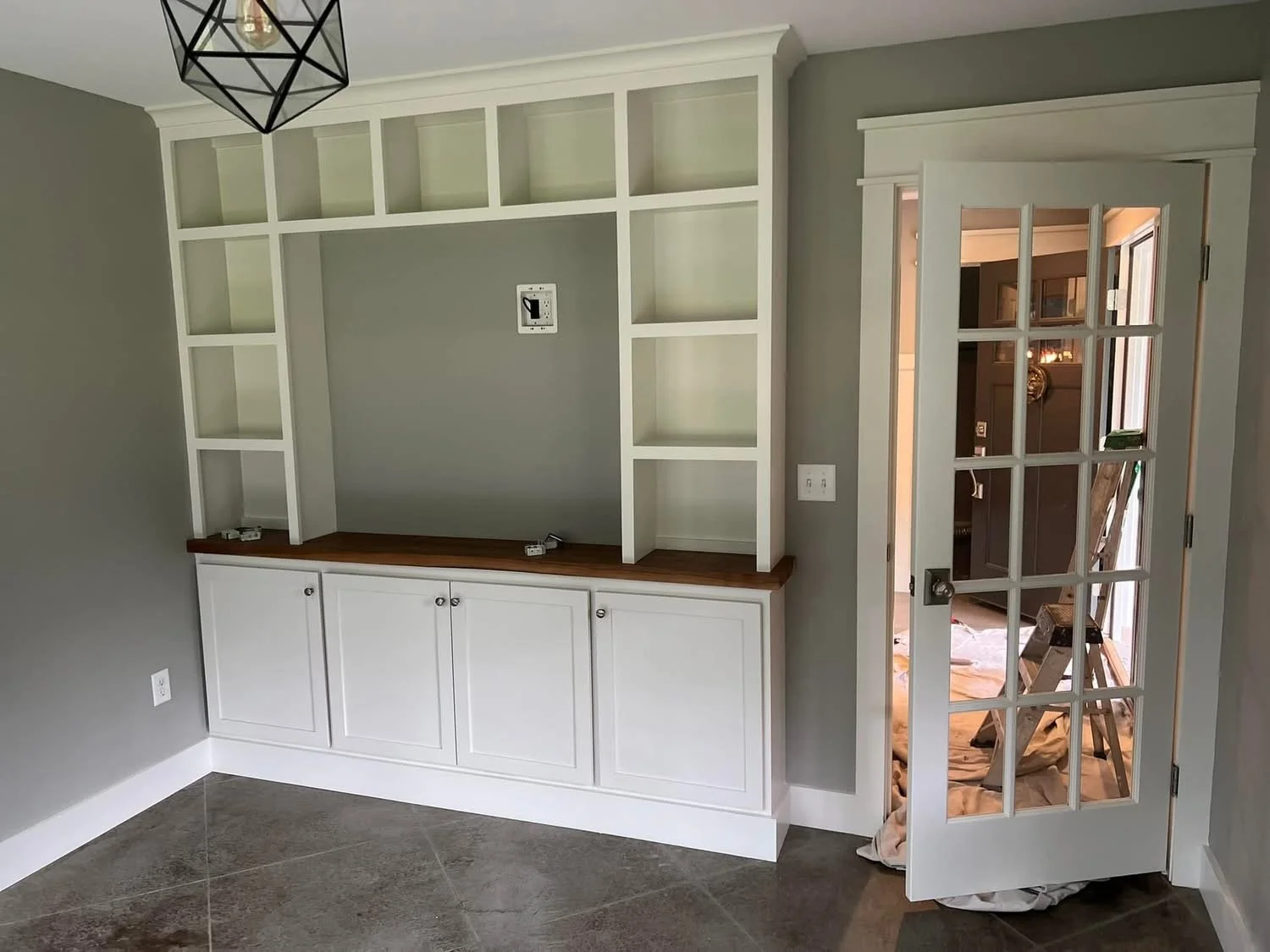 Empty built-in white cabinetry with open shelves and a wooden countertop, situated next to a glass-paneled door leading to another room, in a gray-painted wall.