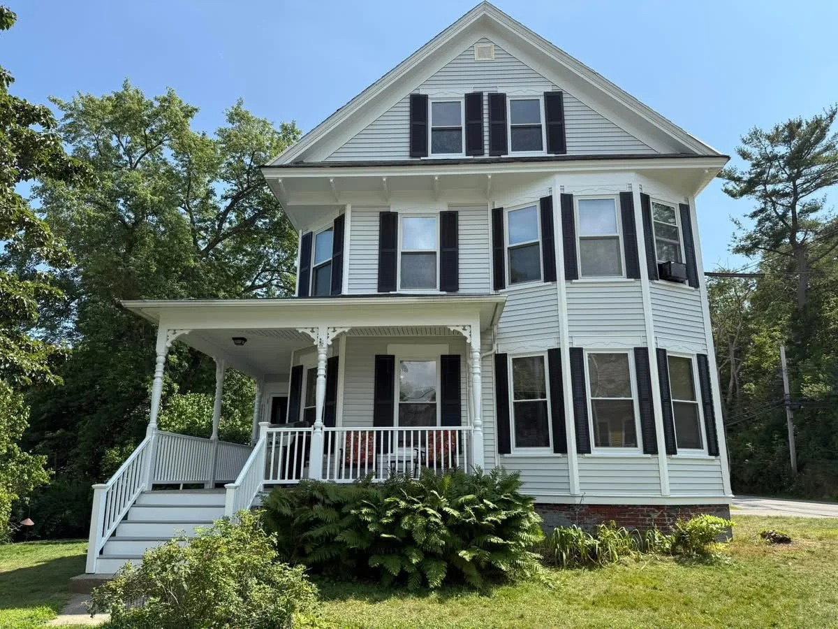 Large white three-story house with black shutters, a front porch with decorative trim, and surrounded by trees and greenery.