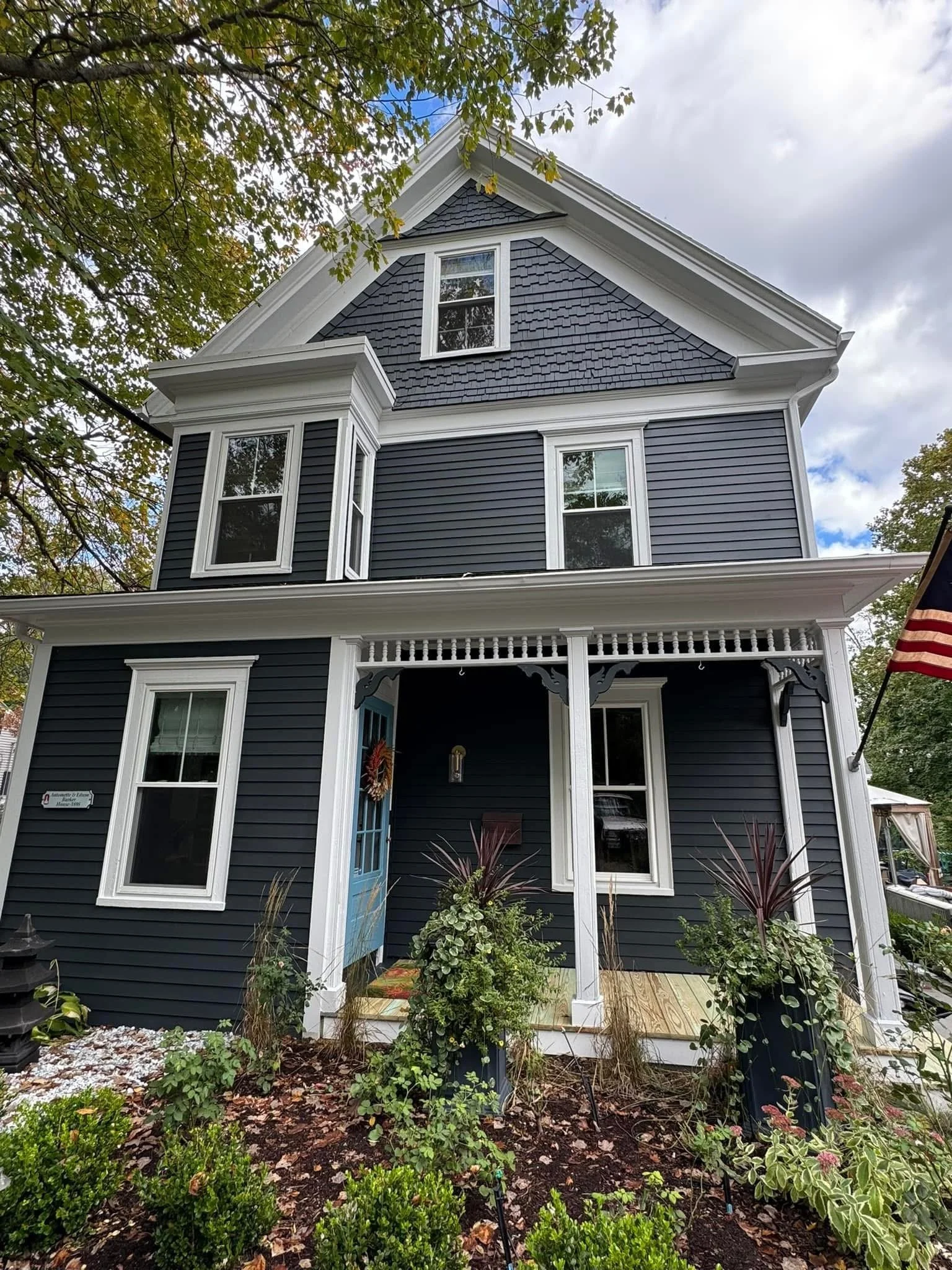A three-story house with dark blue siding, white trim, and multiple large windows. The front porch has a light blue door and is decorated with plants and a wreath. An American flag is hanging on the right side of the house. Trees and a cloudy sky are visible in the background.