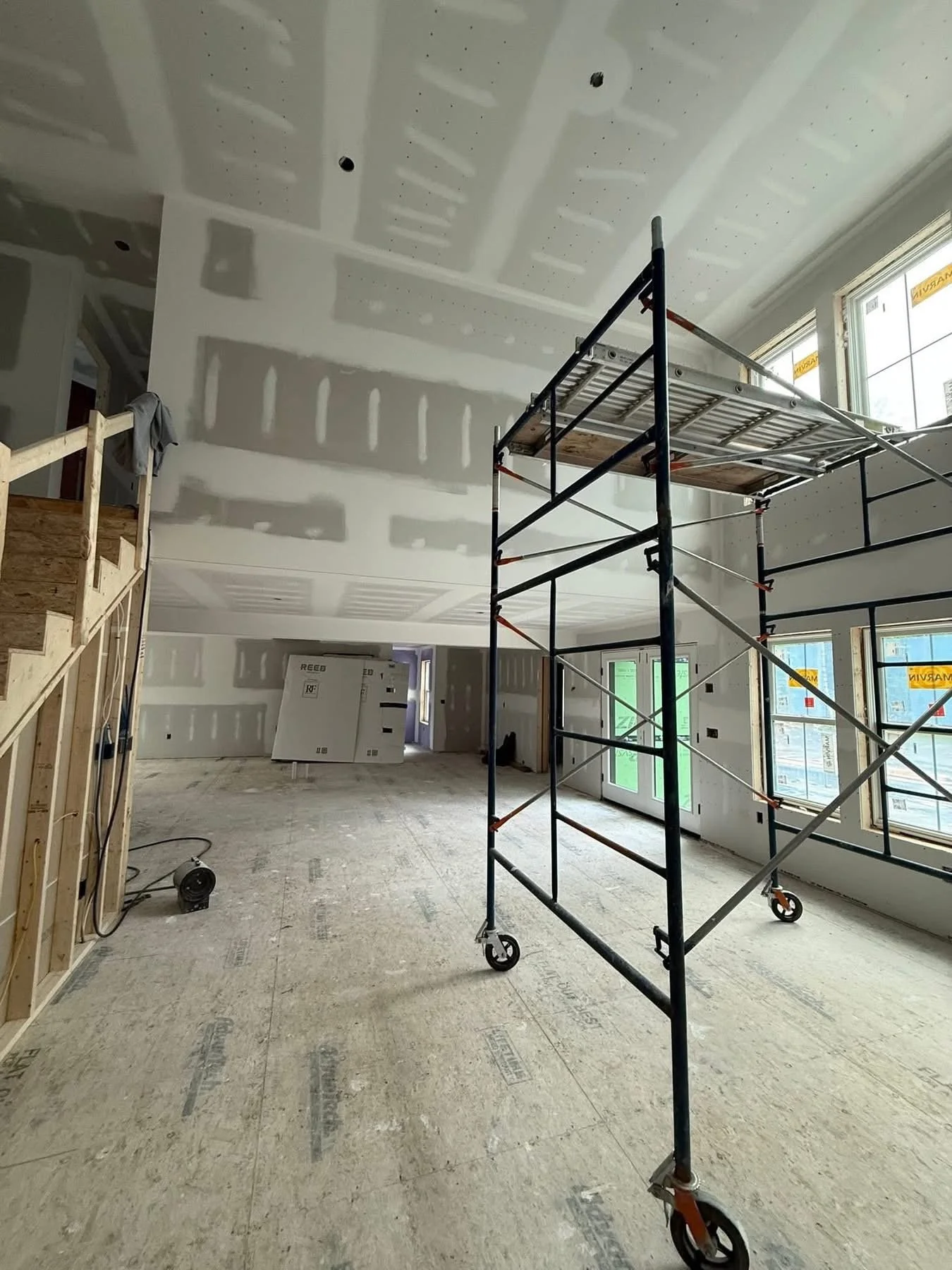 Interior of a house under construction with drywall in progress, scaffolding in the center, and large windows providing natural light.
