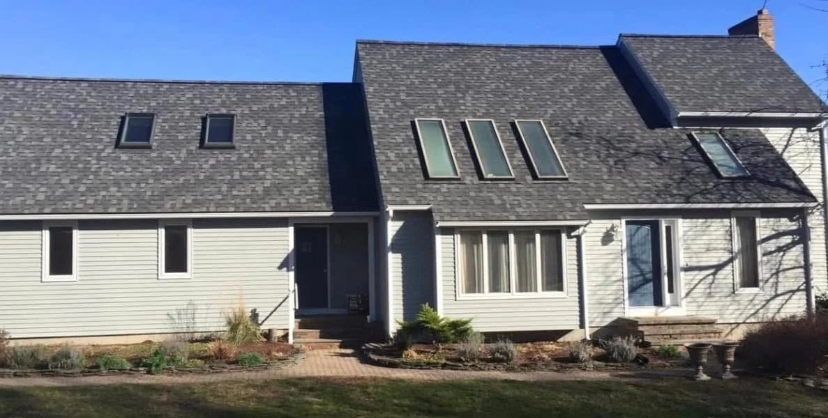 Front view of a two-story house with a grey shingled roof, white siding, and multiple windows, including skylights and a glass door, with a garden and steps leading to the entrance.