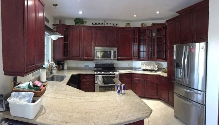 Kitchen with red wooden cabinets, stainless steel refrigerator, microwave, and oven. Beige countertops, a sink beneath a window, and various kitchen items on the counters.