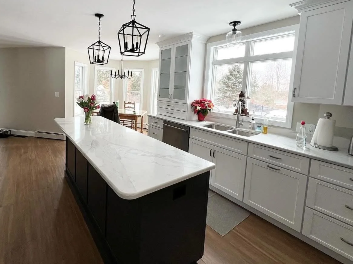 Modern kitchen with white cabinets, large window, marble island, and wooden floors, decorated with flowers and small appliances.