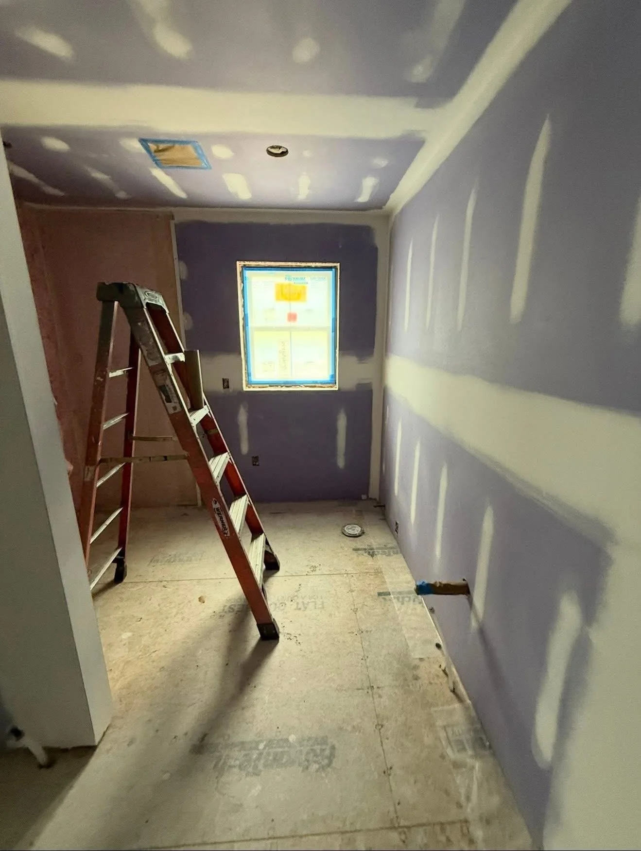 An unfinished room under construction with drywall installed and tape on the seams. A step ladder is centered in the room, and an opening in the floor indicates plumbing work. A window at the end of the room shows a screen with construction plans.