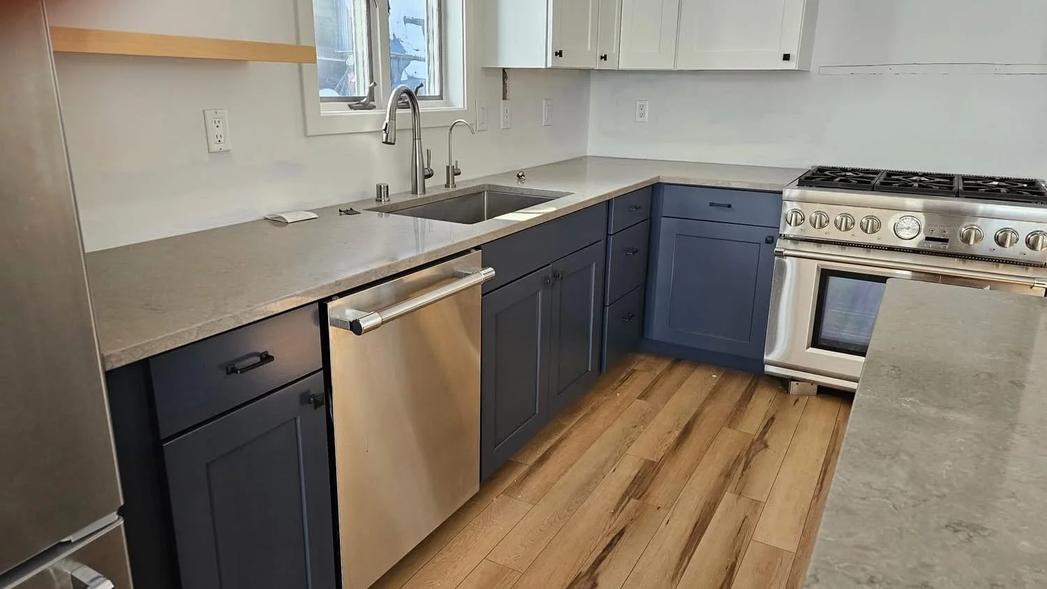 Modern kitchen with white upper cabinets, blue lower cabinets, gray countertops, a stainless steel dishwasher, a stainless steel oven with stove, and hardwood flooring. There is a window above the sink.