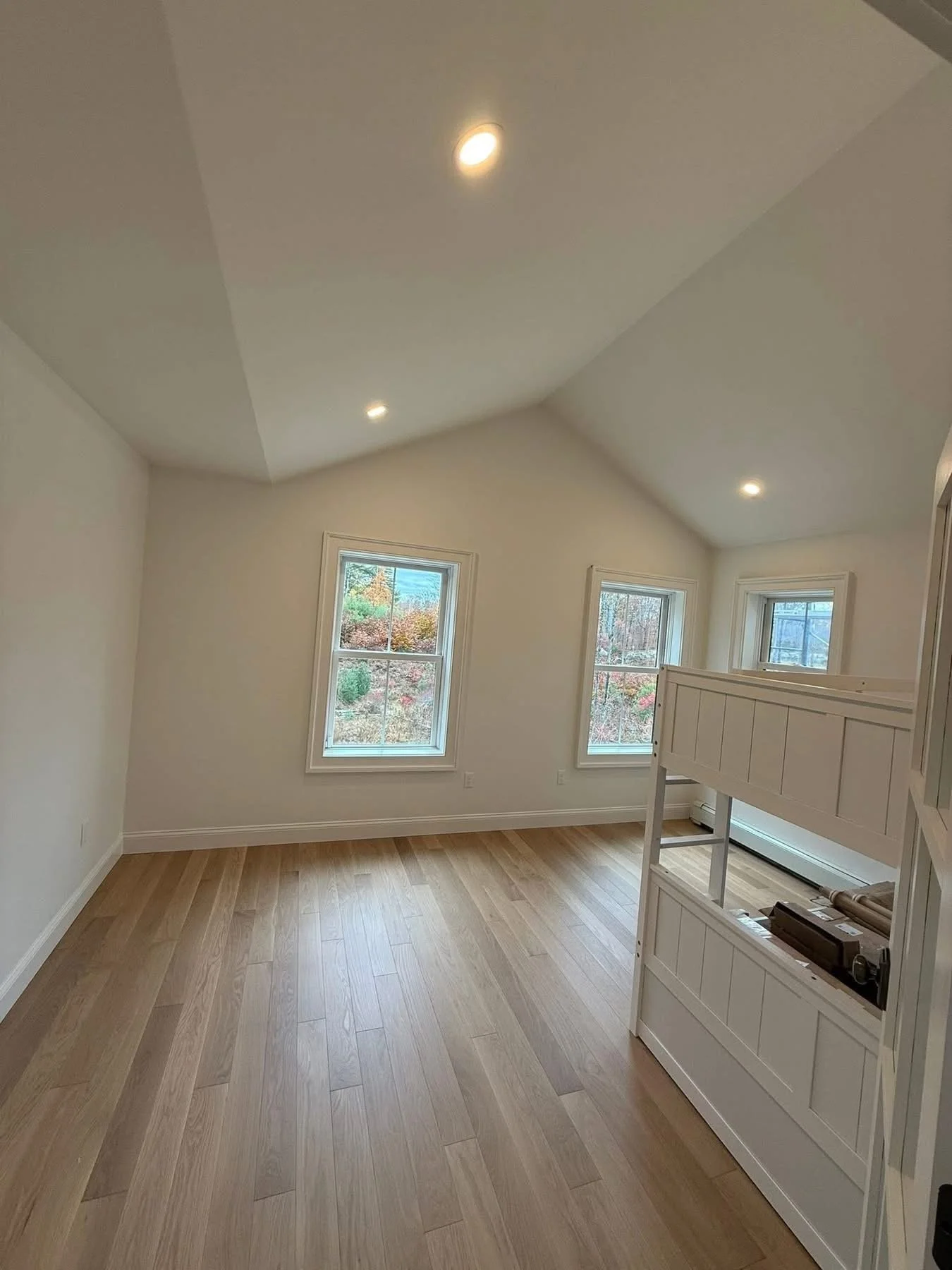 Empty room with beige wooden flooring, white walls, three windows showing outdoor scenery, and ceiling spotlights. A white built-in structure partially blocks the right side.