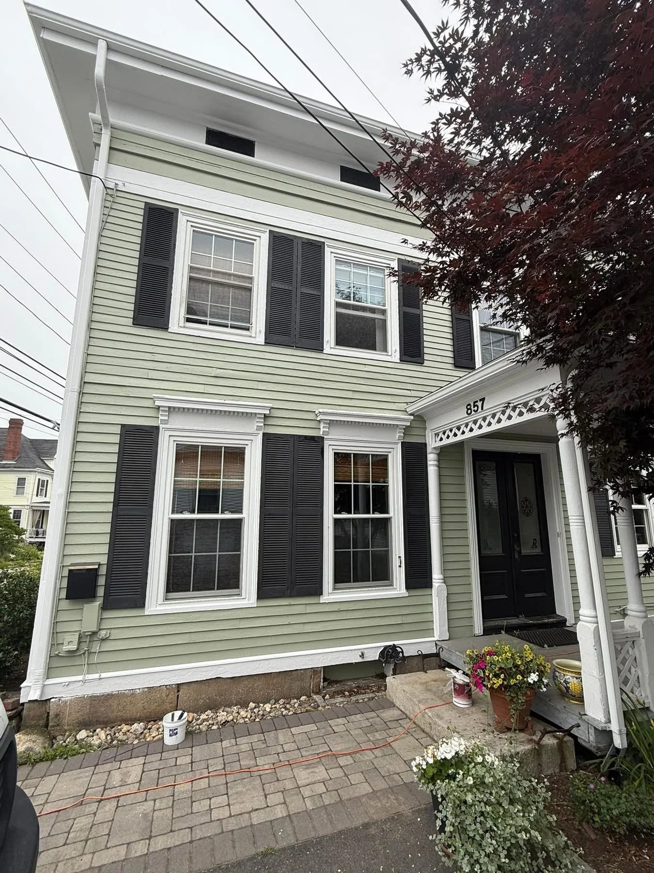 Front of a three-story house with light green siding, black shutters, white trim, and a small porch with decorative white trim. There are two potted plants on the porch and a paved pathway leading to the front door.
