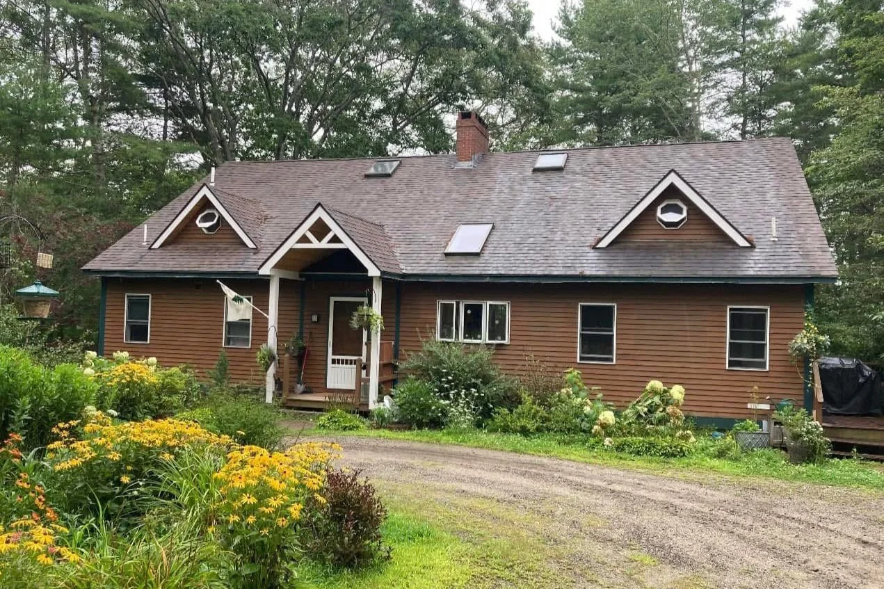 A brown house with a front porch, surrounded by green plants and flowers, trees in the background, and a dirt driveway in front.