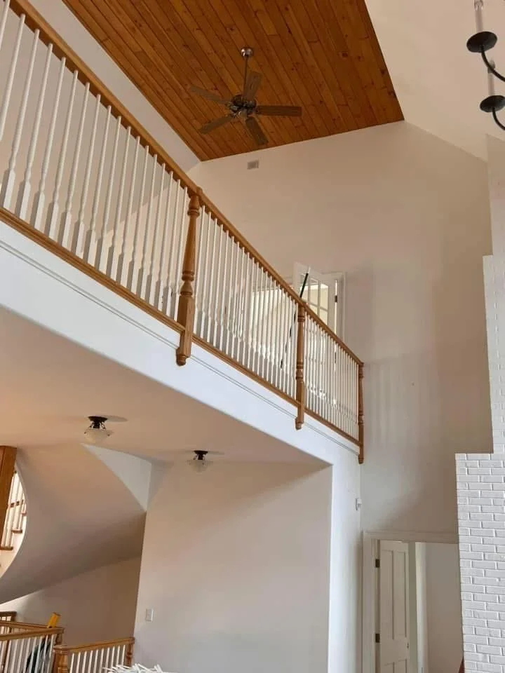Interior view of a two-story house with a high vaulted ceiling, white walls, wooden ceiling panel, ceiling fan, and a wooden railing overlooking the lower level.