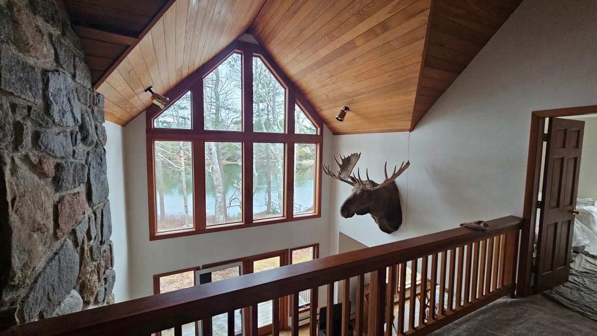 Interior view of a house with a large window displaying trees and water outside, a mounted moose head on the wall, and a wooden railing.