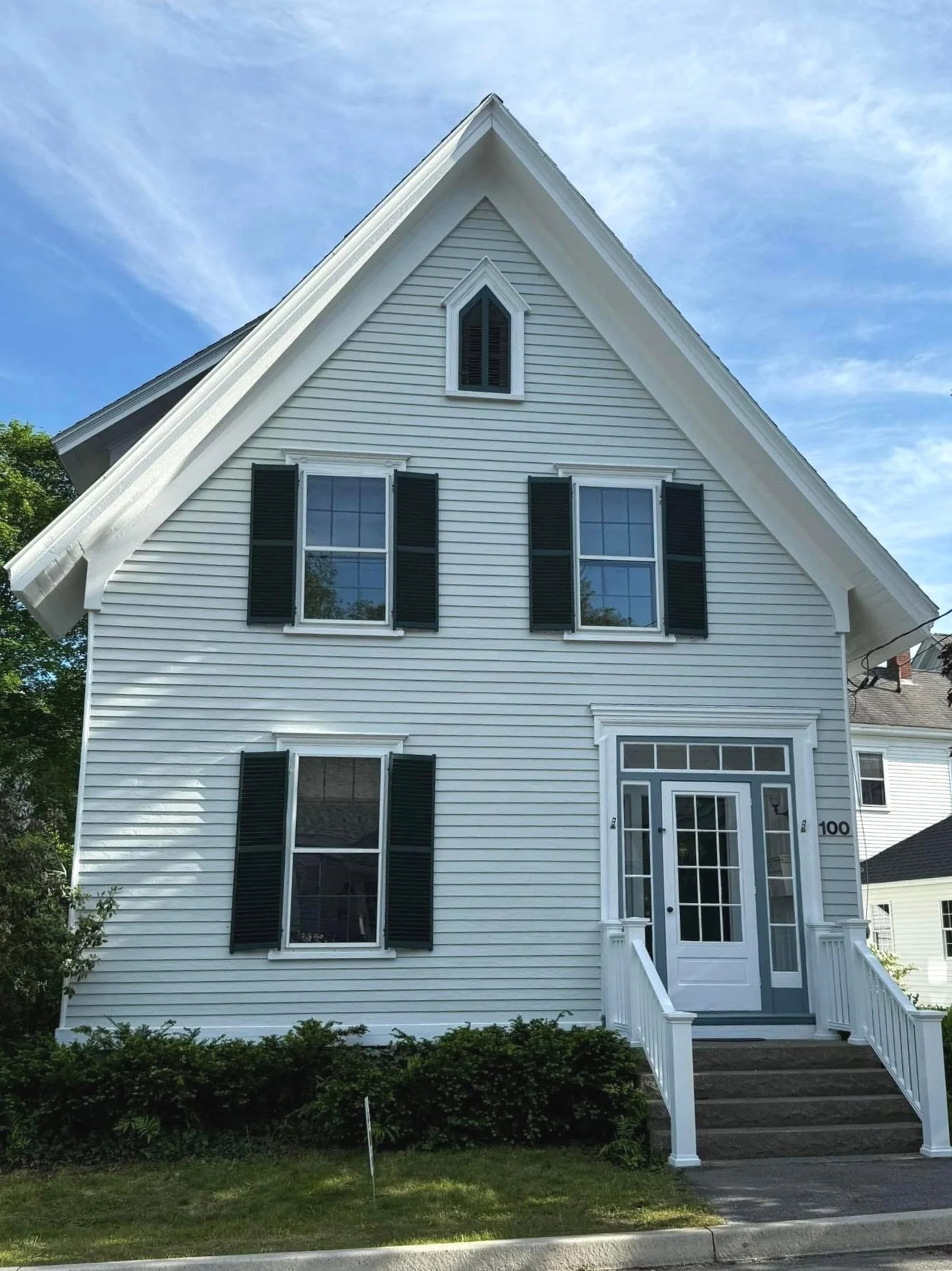 A white house with black shutters, a front porch, and a steep gabled roof, under a partly cloudy sky.