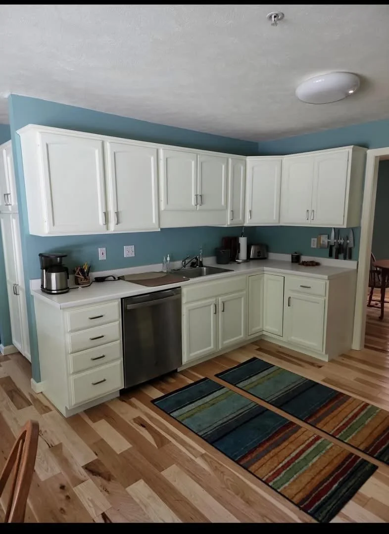 Kitchen with white cabinets, teal wall, wooden floor, and colorful striped rugs. Countertop appliances include a coffee maker, toaster, and knife holder.