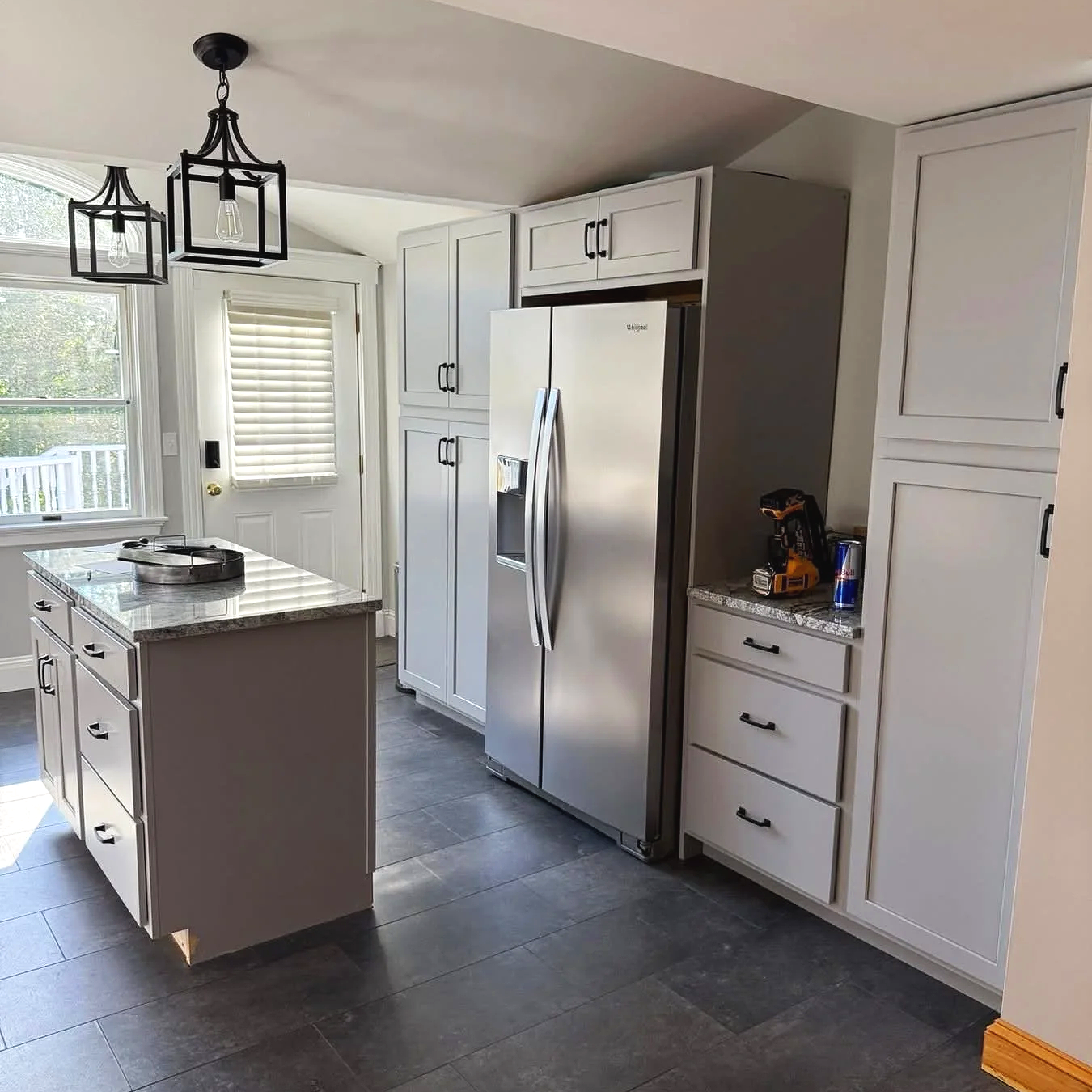 Modern kitchen with white cabinets, stainless steel refrigerator, granite countertop island, dark tile flooring, and two pendant lights.