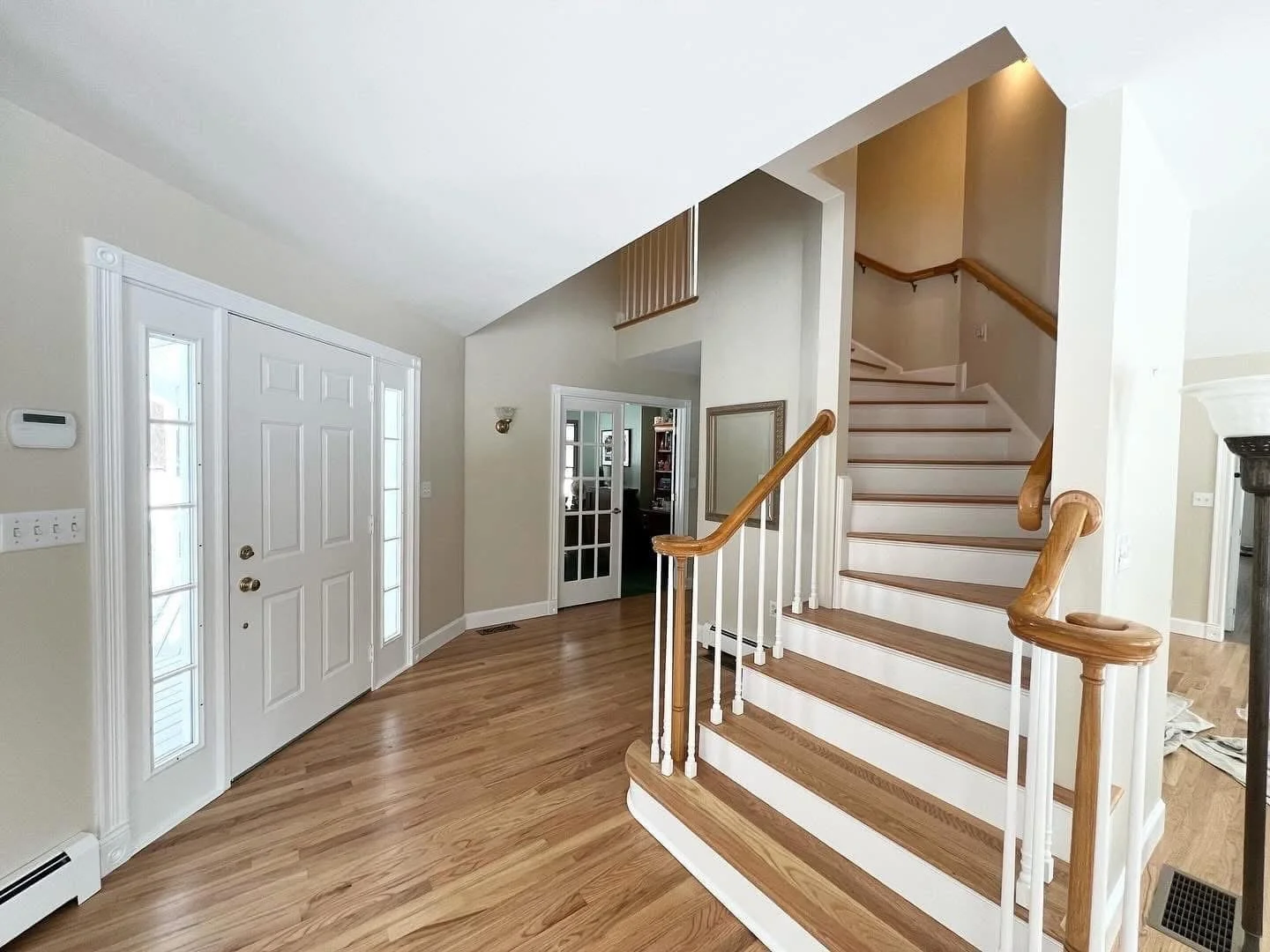 Interior of a house with a staircase, hardwood floors, a front door with sidelights, and a doorway leading to another room.