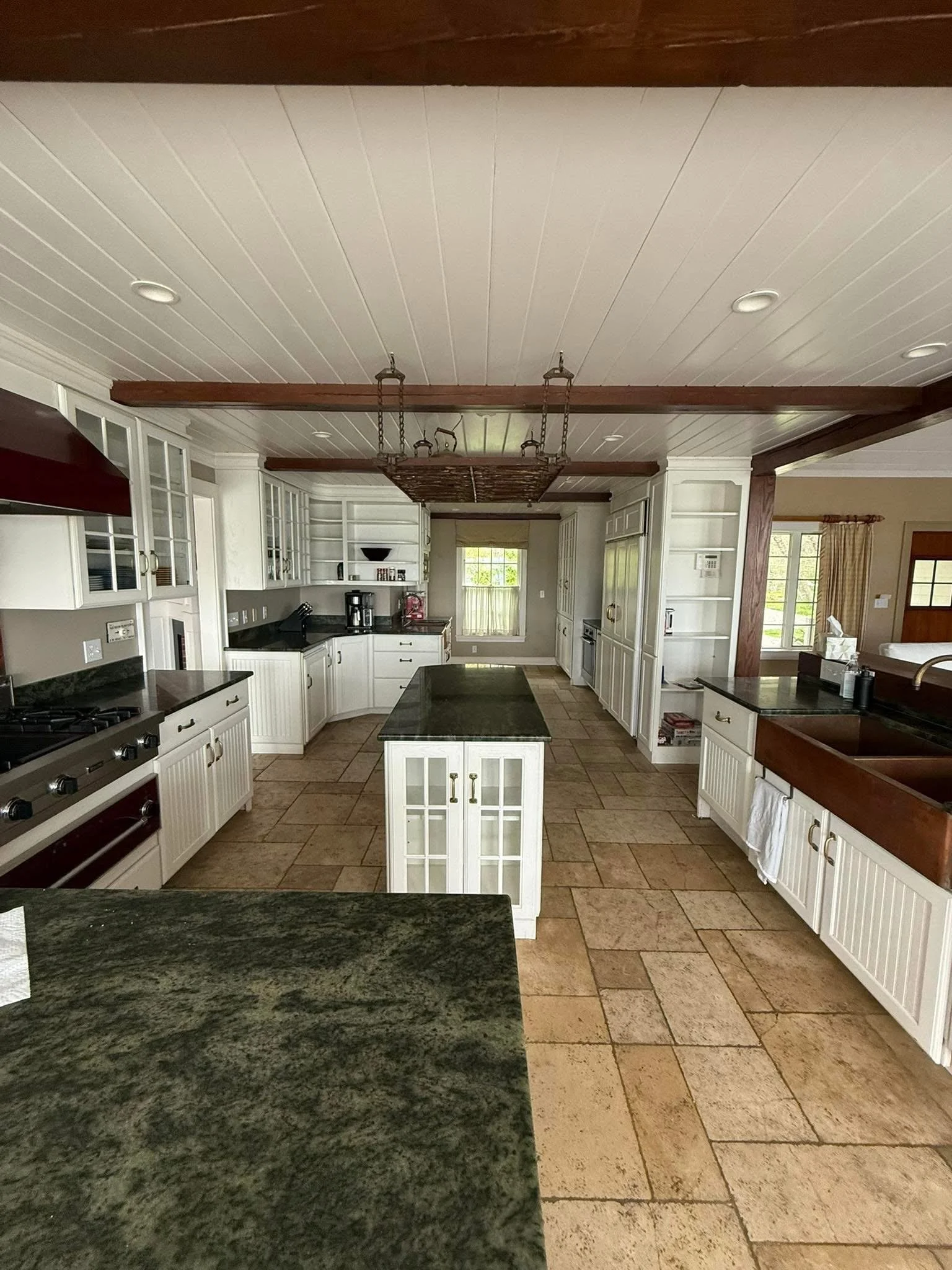 Spacious kitchen with white cabinets, black countertops, a central island, and a copper farmhouse sink. Tiled floor and large windows providing natural light.