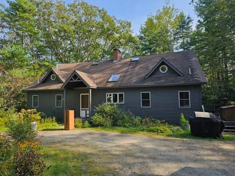A dark gray house with a pitched roof, surrounded by trees and greenery, with a gravel driveway in front and outdoor furniture to the side.