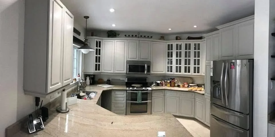 Kitchen with white cabinets, stainless steel refrigerator, oven, microwave, and beige countertops. The counter has various food items and kitchen tools.