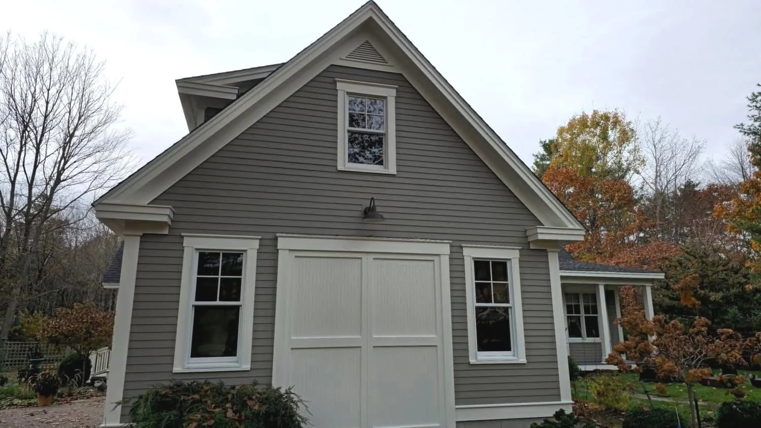 A gray house with white trim, two windows on the lower level, a window and a vent on the upper level, surrounded by trees with fall foliage.